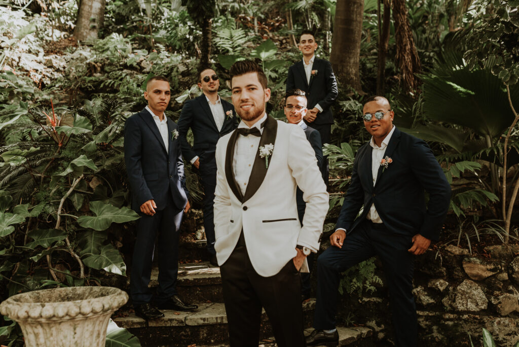 Novio y amigos posan para la cámara  en un jardín antes de la ceremonia de bodas. Novio vestido de blanco y negro al centro, sonríe a la cámara. Boda en La Habana, Cuba