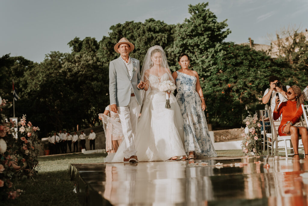 Novia entrando a su ceremonia de bodas acompañada por sus padres. Boda de jardín. Boda de destino en La Habana, Cuba.