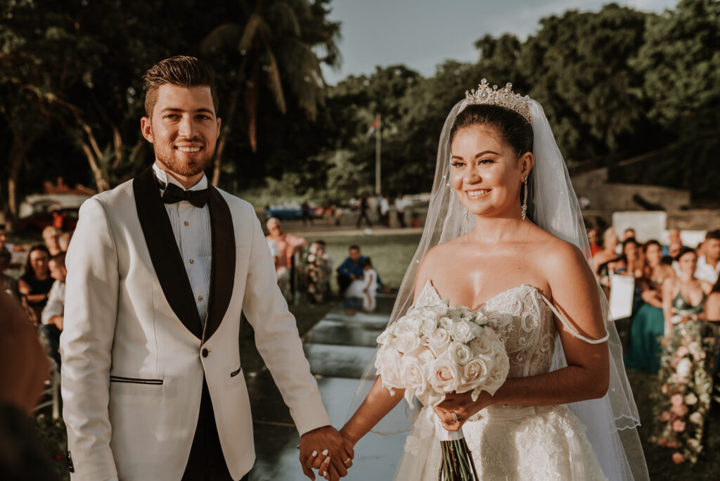 Novios sonríen a la cámara durante su ceremonia de bodas. Novia con vestido blanco, velo y diadema, sostiene ramo de rosas blancas en sus manos. Novio de blanco y negro. Boda en La Habana, Cuba.